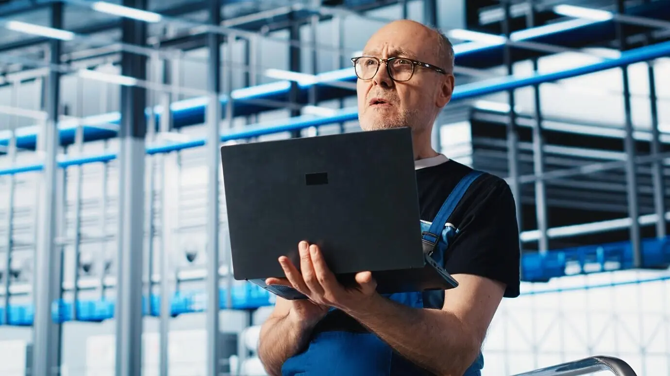 Ältere Ingenieurin/älterer Ingenieur nutzt in einer Smart Factory einen Laptop, um automatisierte Systeme zu testen.